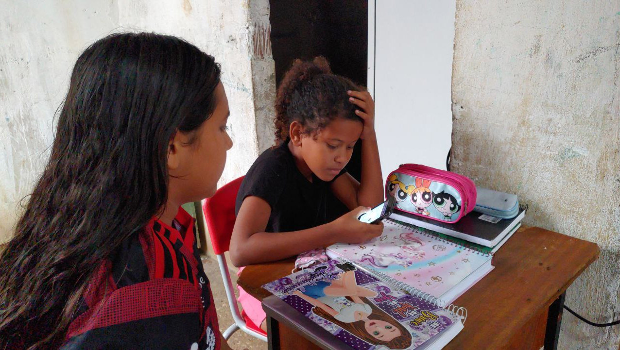 Imagem 1 -  Menina de 9 anos divide celular com o irmão e uma amiga para conseguirem estudar. Maria Mel é aluna do quarto ano do ensino fundamental da Escola Municipal João Niceras de Morais, no Sítio Pedra Branca. Desde 2020, início da pandemia do coronavírus, a menina tem acompanhado as aulas por um pequeno celular doado pela professora, através do aplicativo whatsapp, pois o aparelho não suporta o download do google meet (aplicativo de reuniões por onde as aulas remotas estão sendo ministradas). O equipamento ainda é compartilhado com a amiga Letícia Aparecida, de 12 anos, que cursa a mesma série, e com o irmão José Messias, de 8 anos, que cursa o segundo ano do ensino fundamental.